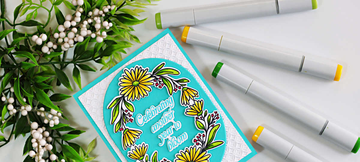Close-up of a handmade birthday card featuring the Simon Says Stamp Daisy Frame, colored with yellow and green Copic markers. The floral frame surrounds the sentiment, "Celebrating another year to bloom," on an Audrey Blue oval, layered over a white panel embossed with the Woven Rings Embossing Folder. Copic markers and greenery frame the card in a bright, clean flat lay.