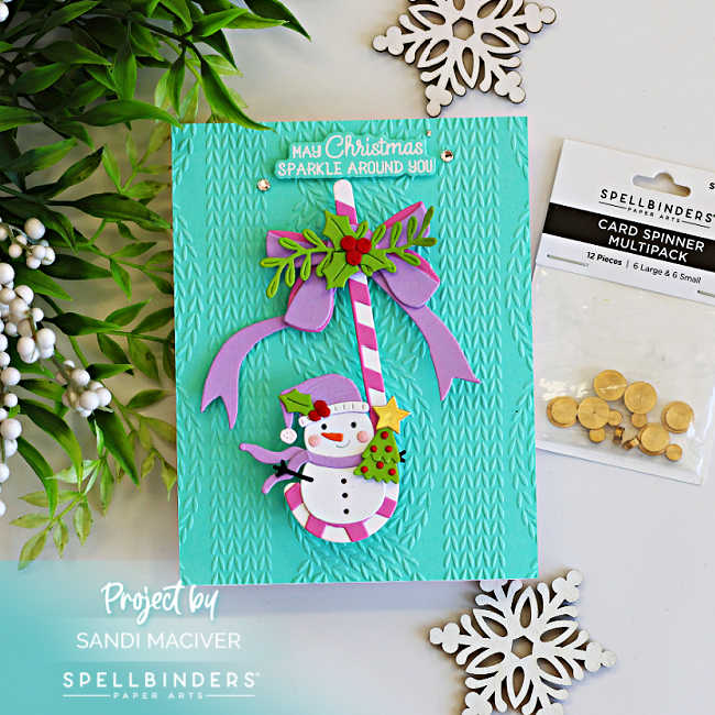 Interactive Christmas card featuring a cheerful snowman swinging from a pink and white candy cane on a turquoise knit embossed background, with the Spellbinders Card Spinner shown beside the finished card.