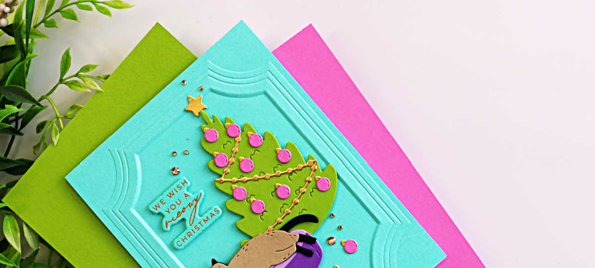 Close-up of a handmade Christmas card featuring a die-cut cat sleeping in a purple pet bed beside a decorated Christmas tree on a turquoise panel embossed with the A2 Ornate Molded Frame 3D Embossing Folder. The card includes a "We Wish You a Meowy Christmas" sentiment and is accented with gold embellishments and pink ornaments.