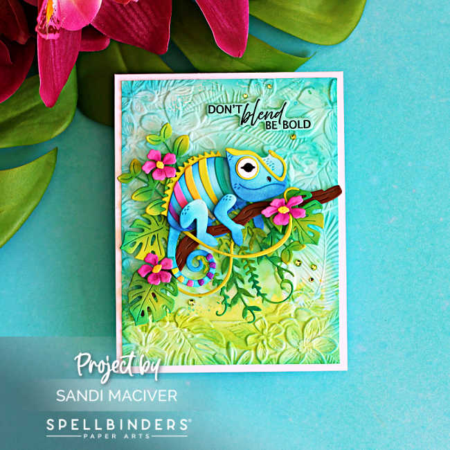 Handmade tropical card featuring the Spellbinders Huey Chameleon die perched on a branch with layered jungle leaves and bright pink flowers over an embossed aqua background. The sentiment reads, "Don't blend, be bold."
