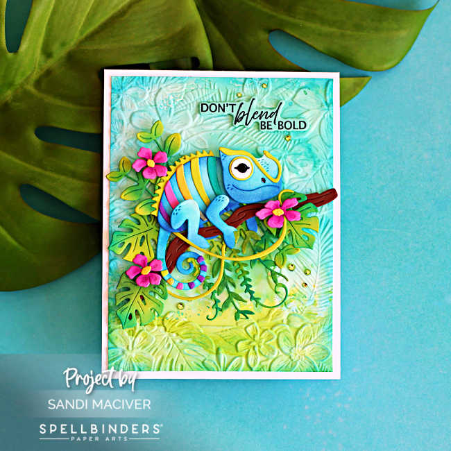 Close-up of a handmade tropical card featuring the Spellbinders Huey Chameleon die on a tree branch surrounded by layered jungle leaves and bright pink flowers. The ink-blended, embossed background creates a lush tropical scene, with the sentiment, "Don't blend, be bold."