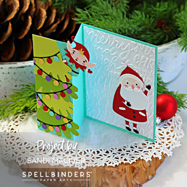 Angled view of a handmade Merry Shuffle Single Z-Fold Christmas card standing open. The front panel features a layered green Christmas tree decorated with colorful holiday lights, while a playful elf peeks from behind the fold. The inside panel showcases a die-cut Santa on a white embossed script background with a turquoise card base. The card is displayed on a rustic wood slice with evergreen branches, a pinecone, and a red Christmas ornament.
