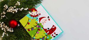 op-down view of a Merry Shuffle Single Z-Fold Christmas card featuring a layered green Christmas tree decorated with colorful lights, a playful elf peeking from behind the fold, and a cheerful Santa on an embossed white background. The card is displayed beside festive greenery and a red Christmas ornament.