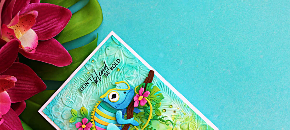 Colorful handmade tropical card featuring the Spellbinders Huey Chameleon die perched on a branch with layered jungle leaves and pink flowers against an embossed aqua background. The sentiment reads, "Don't blend, be bold."
