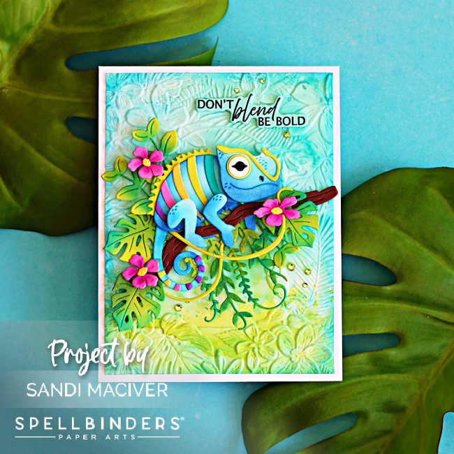 Handmade tropical greeting card featuring the Spellbinders Huey Chameleon die perched on a branch surrounded by layered jungle leaves and pink flowers over an ink-blended, embossed Tropicana background. The sentiment reads, "Don't blend, be bold."