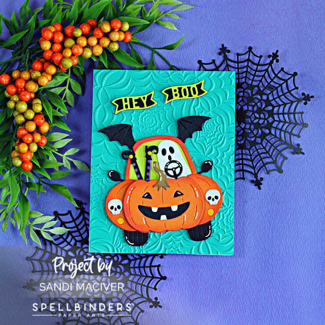 Handmade Halloween card featuring the Spellbinders Boo Mobile Large Die of the Month. A cute ghost drives a pumpkin-shaped car with bat wings and skull headlights on a teal panel embossed with the Spooky Web embossing folder. The card is displayed on a purple background with black spider webs and autumn berry branches.