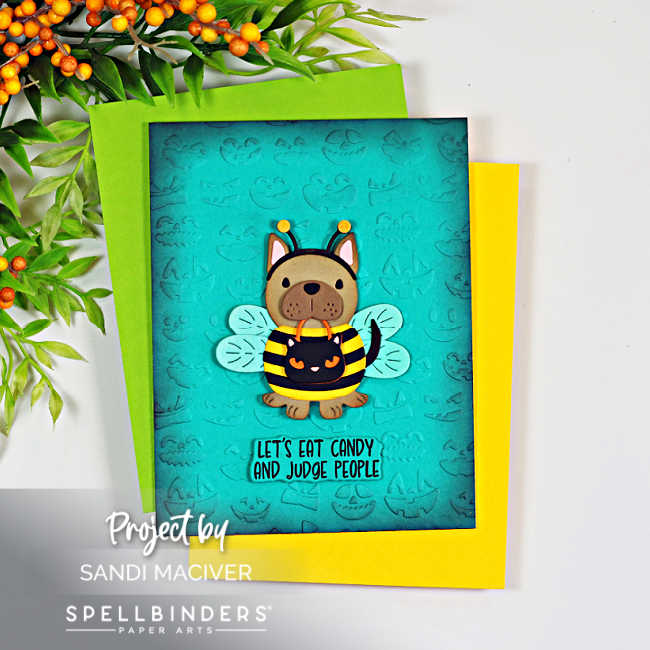 Handmade Halloween card featuring the Spellbinders Buzzy Pup-Bee die set. A cute puppy dressed as a bumblebee with translucent wings stands on a teal ink-blended background embossed with Halloween faces. The playful sentiment reads, "Let's Eat Candy and Judge People," and the card is layered over bright green and yellow cardstock with autumn berry stems in the background.