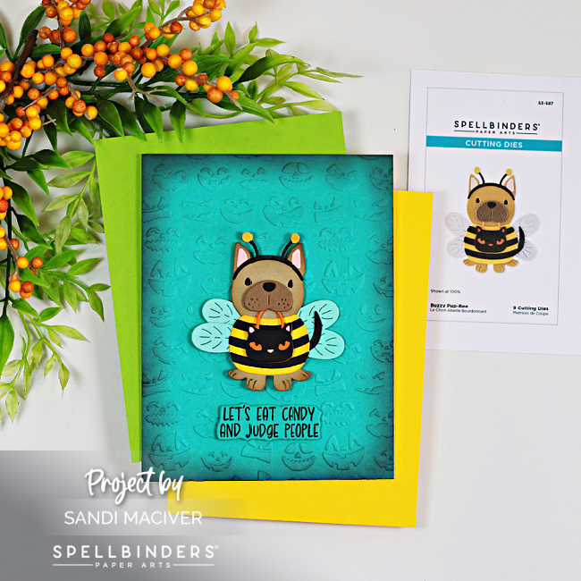 Handmade Halloween card featuring the Spellbinders Buzzy Pup-Bee die set. A cute puppy dressed as a bumblebee with translucent wings stands on a teal ink-blended background embossed with Halloween faces. The playful sentiment reads, "Let's Eat Candy and Judge People," and the card is layered over bright green and yellow cardstock with autumn berry stems in the background.