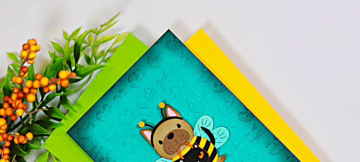 Close-up of a handmade Halloween card featuring the Spellbinders Buzzy Pup-Bee die set. A cute puppy dressed as a bumblebee is showcased on a teal ink-blended background embossed with Halloween icons, layered over bright green and yellow cardstock with autumn berry stems in the corner.