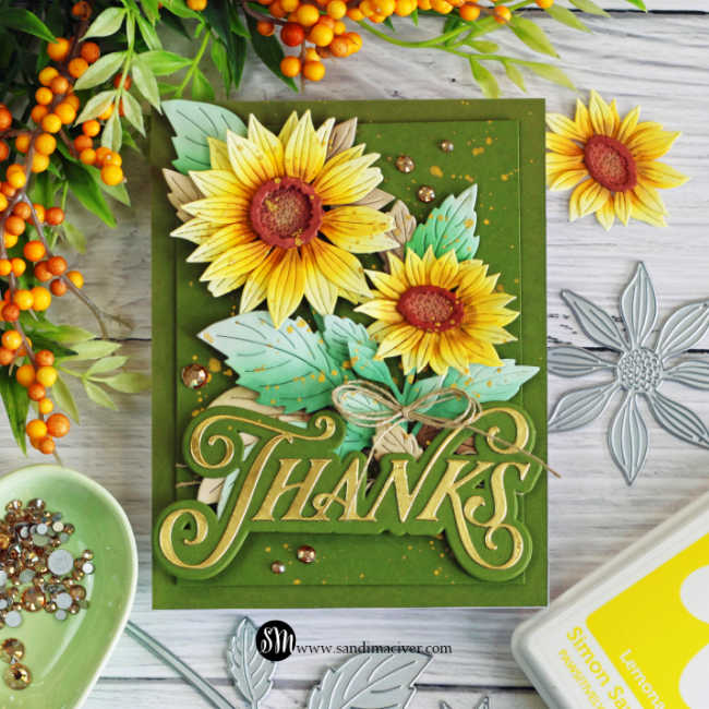 Handmade sunflower thank you card featuring layered yellow die-cut sunflowers, aqua green leaves, an olive green background, gold splatter, gems, twine, and a large gold Thanks sentiment.