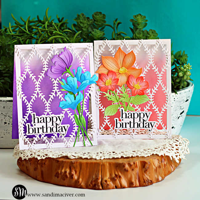 Two handmade open lattice birthday cards displayed on a rustic wood slice with lush greenery in the background. One card features a purple ink-blended background with purple and blue die-cut flowers, while the other showcases a soft pink background with peach and coral flowers. Both cards are finished with intricate white die-cut lattice panels, layered green foliage, and bold black "happy birthday" sentiments.