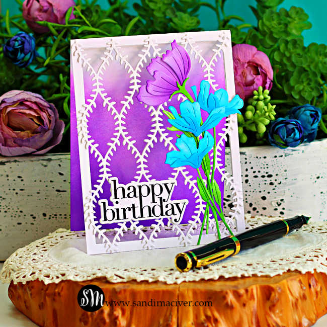 Handmade open lattice birthday card featuring a white die-cut lattice panel over a purple ink-blended background. The card is decorated with a large purple flower and layered blue blooms with green stems and leaves, finished with a bold black "happy birthday" sentiment. The card is displayed on a rustic wood slice with colorful flowers, greenery, and a black fountain pen in the background.