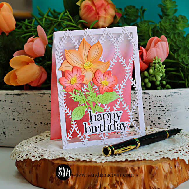 Handmade open lattice birthday card featuring a white die-cut lattice panel over a soft coral and pink ink-blended background. The card is decorated with a large peach flower, two coral blossoms, layered green leaves, and a bold black "happy birthday" sentiment. Displayed on a rustic wood slice with a black fountain pen, the background includes lush greenery and peach flowers.