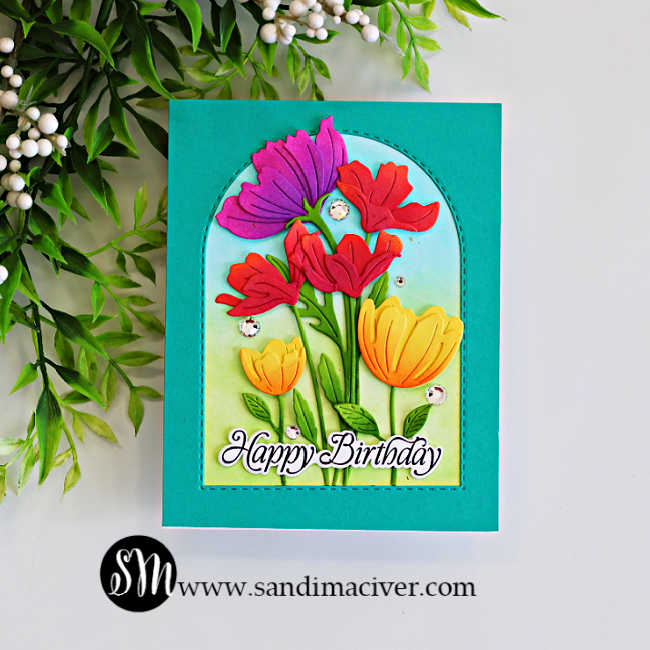 Colorful floral birthday card with ink-blended pink, red and yellow die-cut flowers arranged inside a stitched arch frame on a teal background.
