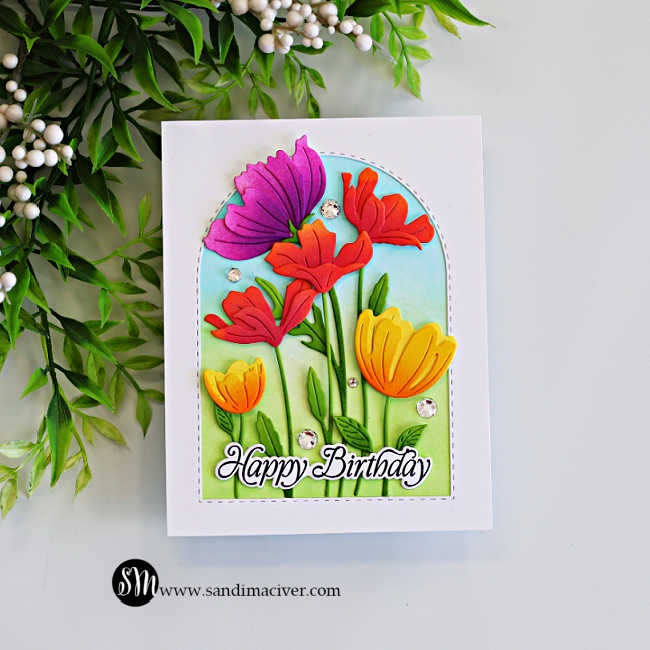 Colorful floral birthday card with ink-blended pink, red and yellow die-cut flowers arranged inside a stitched arch frame on a clean white background.