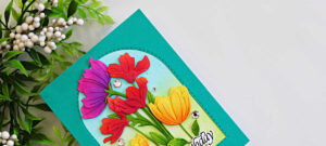 Colorful die-cut flowers arranged inside a stitched arch frame with a teal card background and softly ink-blended blue and green garden backdrop.