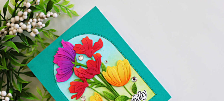Colorful die-cut flowers arranged inside a stitched arch frame with a teal card background and softly ink-blended blue and green garden backdrop.