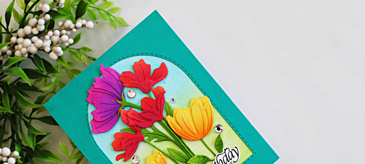 Colorful die-cut flowers arranged inside a stitched arch frame with a teal card background and softly ink-blended blue and green garden backdrop.