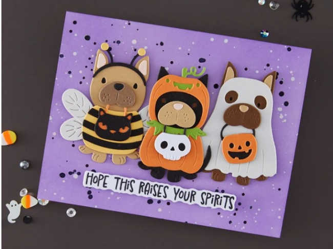 Three adorable Halloween pups created with the Spellbinders Howl-O-Ween Pup Trio Bundle are featured on a purple ink-blended card with black and white paint splatter. The die-cut dogs are dressed as a bumblebee, a pumpkin, and a ghost, with the sentiment, "Hope This Raises Your Spirits," stamped below. Fun Halloween embellishments complete this cute handmade card.
