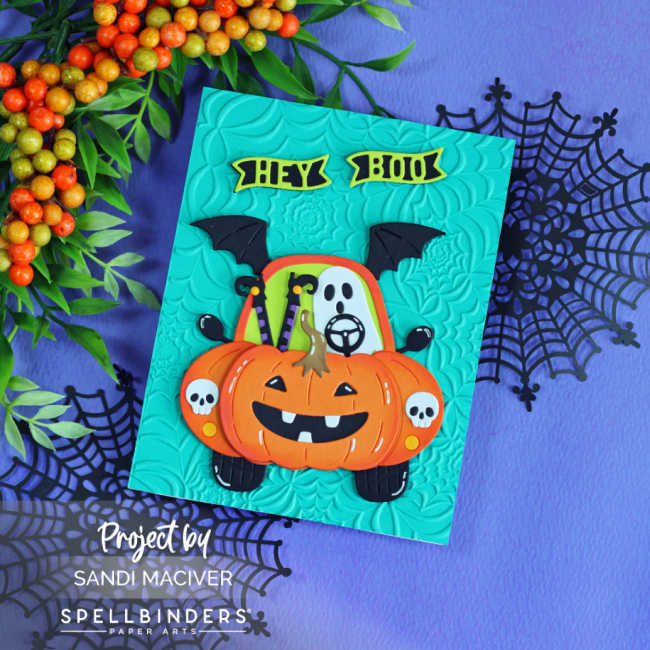 Handmade Halloween card featuring the Spellbinders Boo Mobile Large Die of the Month. A cute ghost drives a pumpkin-shaped car with bat wings and skull headlights on a teal panel embossed with the Spooky Web embossing folder. The card is displayed on a purple background with black spider webs and autumn berry branches.