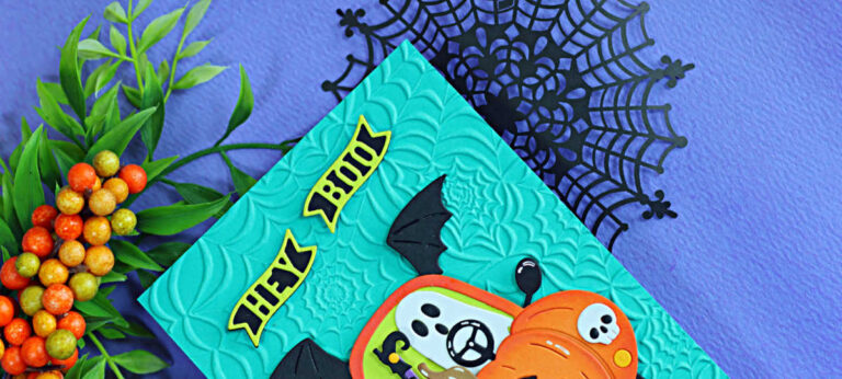 Close-up of a handmade Halloween card featuring the Spellbinders Boo Mobile die set. A smiling ghost drives a pumpkin-shaped car with black bat wings against a teal background embossed with the Spooky Web embossing folder. The card is photographed on a purple background with black spider webs and fall berry branches.