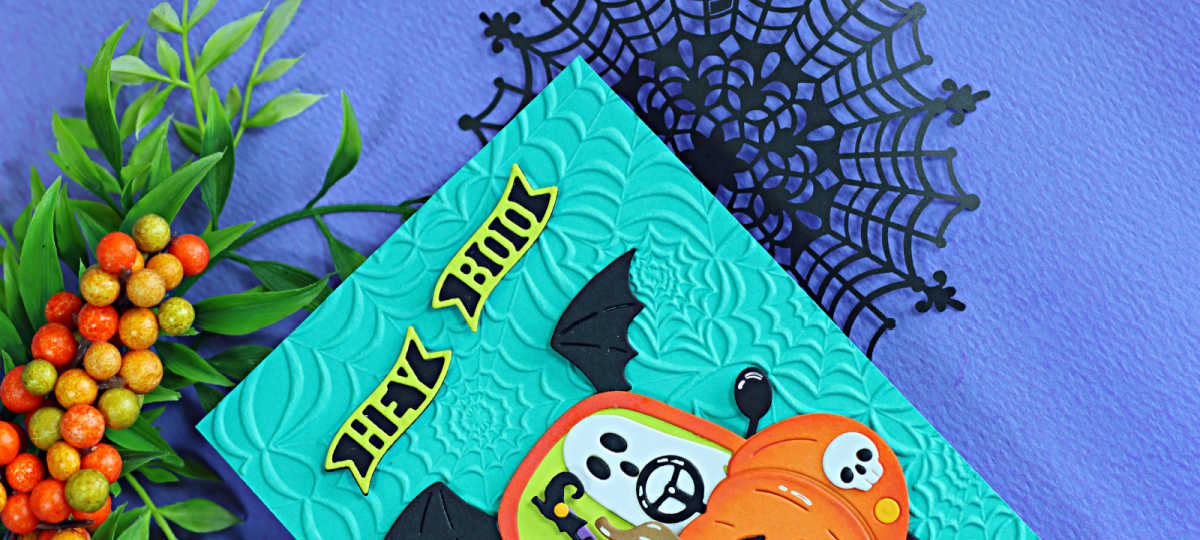 Close-up of a handmade Halloween card featuring the Spellbinders Boo Mobile die set. A smiling ghost drives a pumpkin-shaped car with black bat wings against a teal background embossed with the Spooky Web embossing folder. The card is photographed on a purple background with black spider webs and fall berry branches.