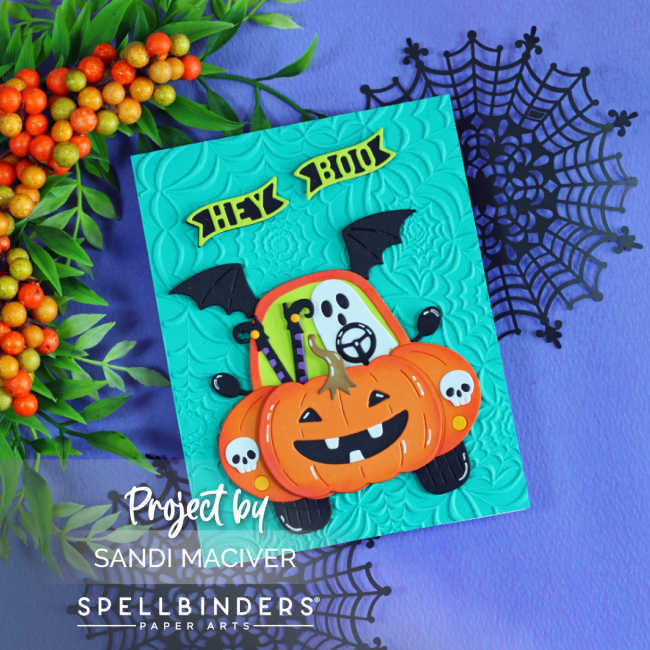Handmade Halloween card featuring the Spellbinders Boo Mobile Large Die of the Month. A cute ghost drives a pumpkin-shaped car with bat wings and skull headlights on a teal panel embossed with the Spooky Web embossing folder. The card is displayed on a purple background with black spider webs and autumn berry branches.