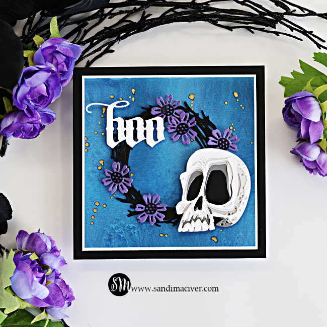 Tim Holtz Halloween card featuring the Spencer Colorize skull with a black foliage wreath, purple flowers, and white Gothic “Boo” sentiment on a textured blue background.