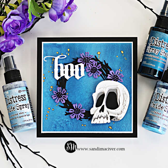 Tim Holtz Halloween card featuring Spencer Colorize, a black foliage wreath with purple flowers, and a white Gothic “Boo” sentiment on a blue Distress Oxide Spray background, staged with Distress Spray Stains.