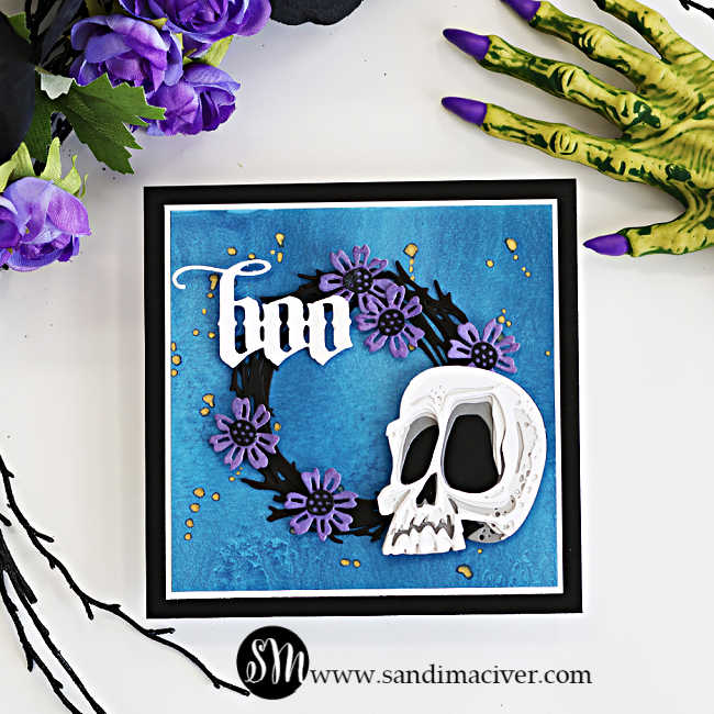 Tim Holtz Halloween card featuring the Spencer Colorize skull, black foliage wreath with purple flowers, and white Gothic “Boo” sentiment on a textured blue background, staged with purple flowers and a creepy green Halloween hand.