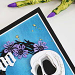 Tim Holtz Halloween card featuring the Spencer Colorize skull, purple flowers on a black foliage wreath, and a white Gothic “Boo” sentiment on a textured blue background, staged with purple flowers and a creepy Halloween hand.
