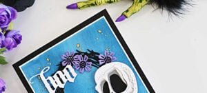 Tim Holtz Halloween card featuring the Spencer Colorize skull, purple flowers on a black foliage wreath, and a white Gothic “Boo” sentiment on a textured blue background, staged with purple flowers and a creepy Halloween hand.