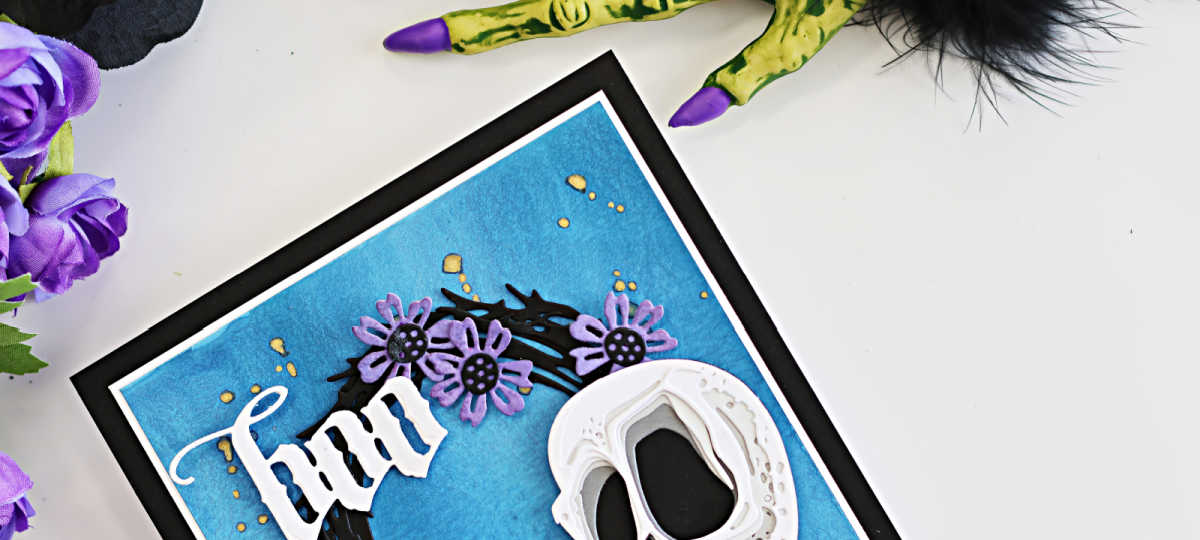 Tim Holtz Halloween card featuring the Spencer Colorize skull, purple flowers on a black foliage wreath, and a white Gothic “Boo” sentiment on a textured blue background, staged with purple flowers and a creepy Halloween hand.