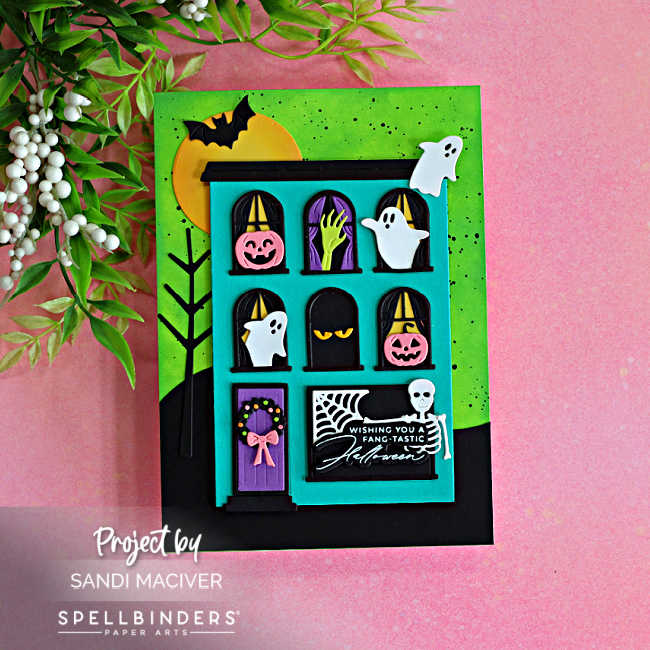 Fang-tastic Halloween card featuring a turquoise haunted house decorated with ghosts, pink pumpkins, a spooky green hand, bat, spiderweb and skeleton on a bright green background.