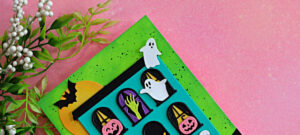 Colorful Halloween card featuring a turquoise haunted house with ghosts, pumpkins, bats and a spooky green hand against an ink-blended green background.