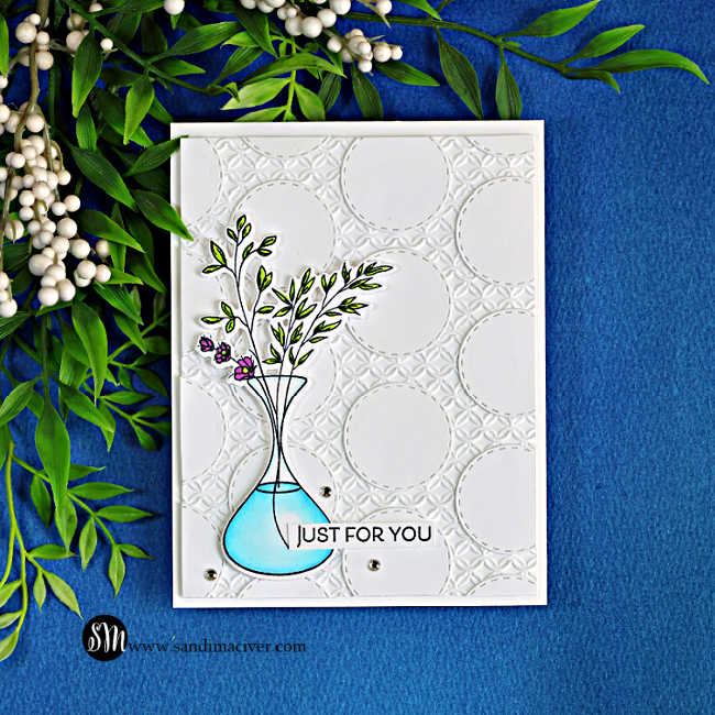 Handmade card featuring a white geometric background created with stitched circle nesting dies, topped with leafy stems and small purple flowers in a turquoise glass vase.