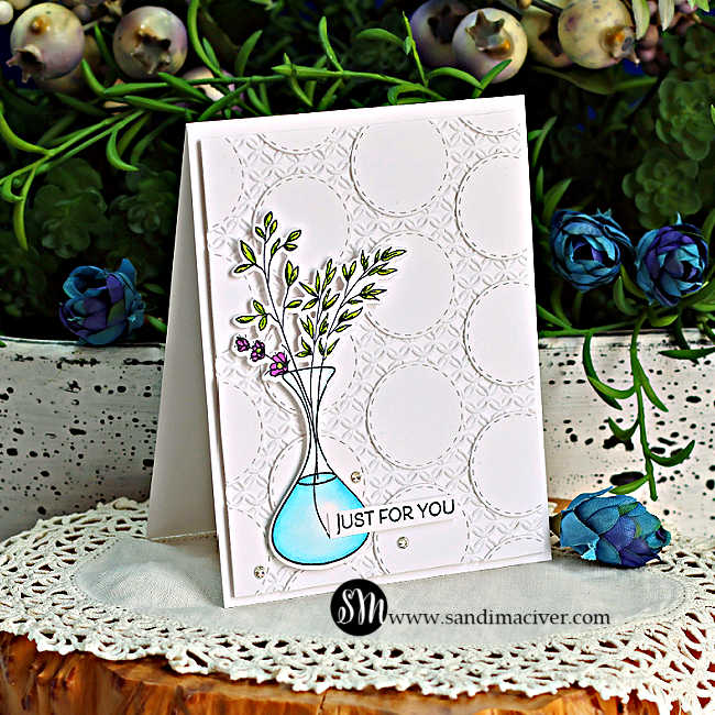 Handmade white card featuring a geometric stitched circle background, accented with leafy stems and tiny purple flowers in a turquoise vase, displayed against lush greenery.
