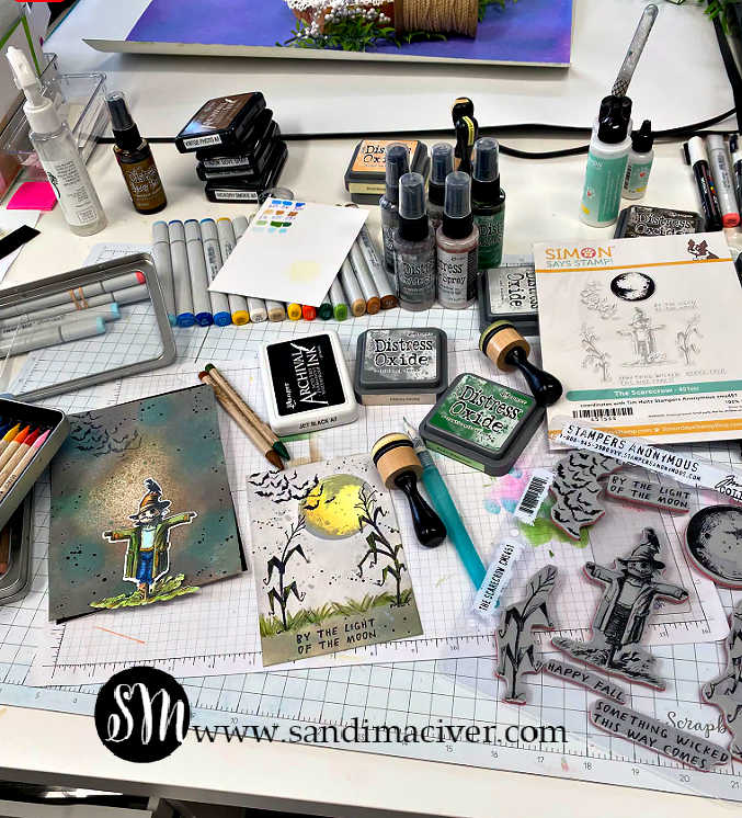 Behind-the-scenes craft desk featuring the Tim Holtz The Scarecrow stamp set and new coordinating dies, Distress Oxide inks, markers, sprays, blending tools, and the Halloween card in progress.