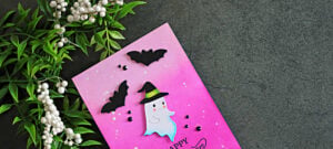 Pink Halloween card featuring a cute white ghost in a witch hat, black die-cut bats, and a bright pink ink-blended background with white splatters.