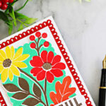 Close-up of a colorful Concord & 9th Incredibly Grateful floral card featuring red and yellow flowers, mint green background, and a decorative red die-cut frame, styled with red berries and a black fountain pen.