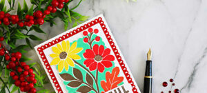 Close-up of a colorful Concord & 9th Incredibly Grateful floral card featuring red and yellow flowers, mint green background, and a decorative red die-cut frame, styled with red berries and a black fountain pen.