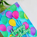 Close-up of a colorful birthday card featuring embossed party balloons in pink, purple, green, yellow, and blue against a turquoise background, finished with a layered Happy Birthday sentiment.