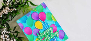 Close-up of a colorful birthday card featuring embossed party balloons in pink, purple, green, yellow, and blue against a turquoise background, finished with a layered Happy Birthday sentiment.