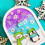 Memory Box Stay Cozy winter card featuring a snowman and penguin on an arch-shaped snowy scene with a purple and turquoise sky and white snowflakes.