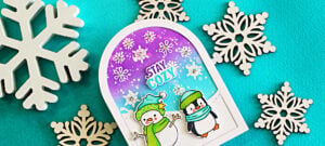 Memory Box Stay Cozy winter card featuring a snowman and penguin on an arch-shaped snowy scene with a purple and turquoise sky and white snowflakes.