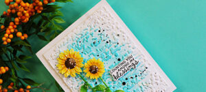 Close-up of a handmade fall card featuring bright yellow die-cut sunflowers against a textured aqua and white background with black and white splatters and a “Counting You Among My Blessings” sentiment.