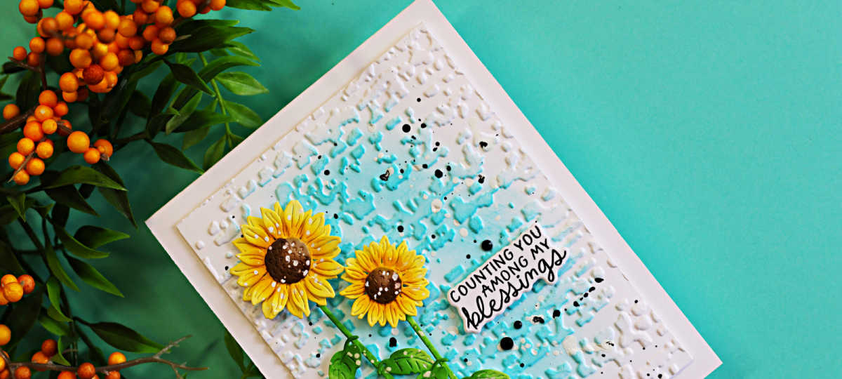 Close-up of a handmade fall card featuring bright yellow die-cut sunflowers against a textured aqua and white background with black and white splatters and a “Counting You Among My Blessings” sentiment.