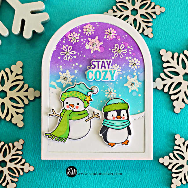 Memory Box Stay Cozy winter card with a snowman and penguin on a snowy arch, set against a purple and turquoise snowflake-filled sky.