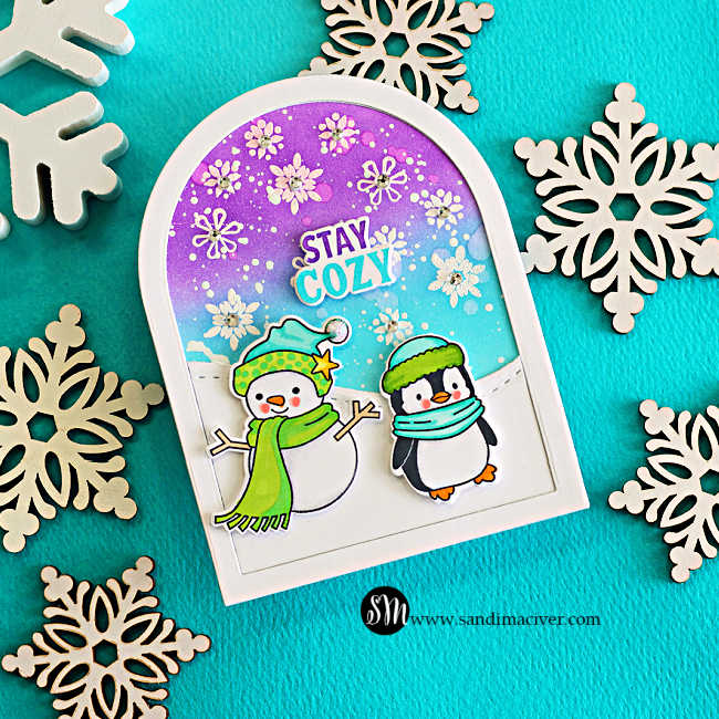Memory Box Stay Cozy winter card with a snowman and penguin on a snowy arch, set against a purple and turquoise snowflake-filled sky.