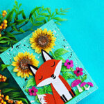 Spellbinders Hello Foxy fall card featuring a die-cut fox surrounded by sunflowers, bright pink flowers and mushrooms on a Waterfall embossed background.